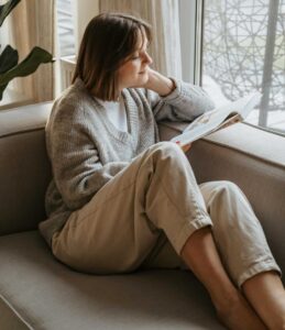 A woman reading a book on a couch near a window, representing relaxation, reflection, and quiet emotional space.